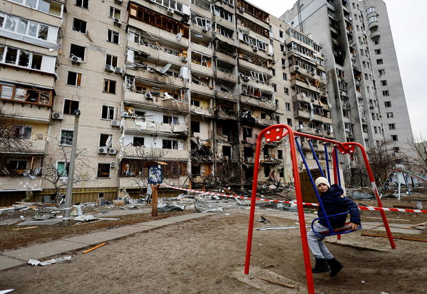 A boy on a red swing set look at camera in front of a bombed out residential building in Ukraine