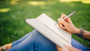 A woman writing in a notebook with a pen while sitting outside, maybe in a park. She is on the grass with the notebook up against her bent knees.